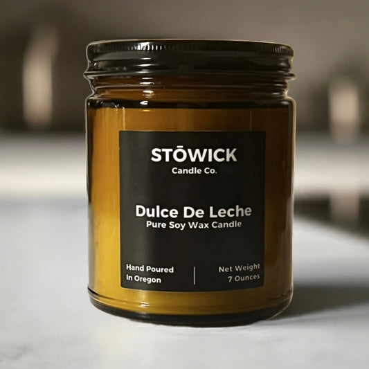 A 7oz soy wax dulce de leche jar candle in an amber jar with a black lid sitting on a white table.