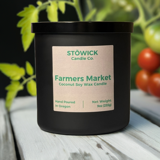 A farmers market scented candle in a 9 ounce black jar with a tan label sitting outside on a white wood table with tomato vines in the background