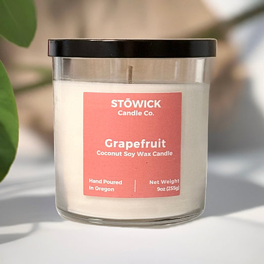A grapefruit scented soy coconut wax candle in a clear jar with a pink label and a black lid on top of a table next to a grapefruit plant