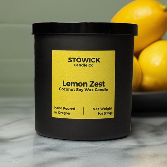 A 9 ounce lemon zest scented soy coconut wax candle in a black matte jar with a yellow label sitting on a kitchen counter with lemons behind it.