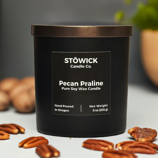 A 9 ounce pecan praline scented soy wax candle in a black with a bronze lid and a black label sitting on a table next to pecans