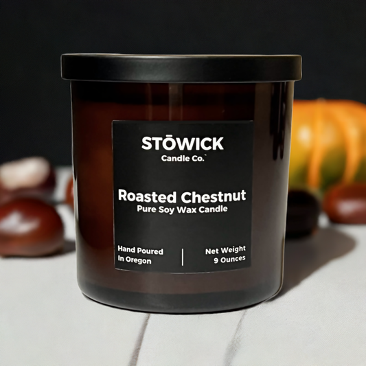 a roasted chestnut soy wax candle in an amber jar with a black lid and label sitting on a white table by chestnuts