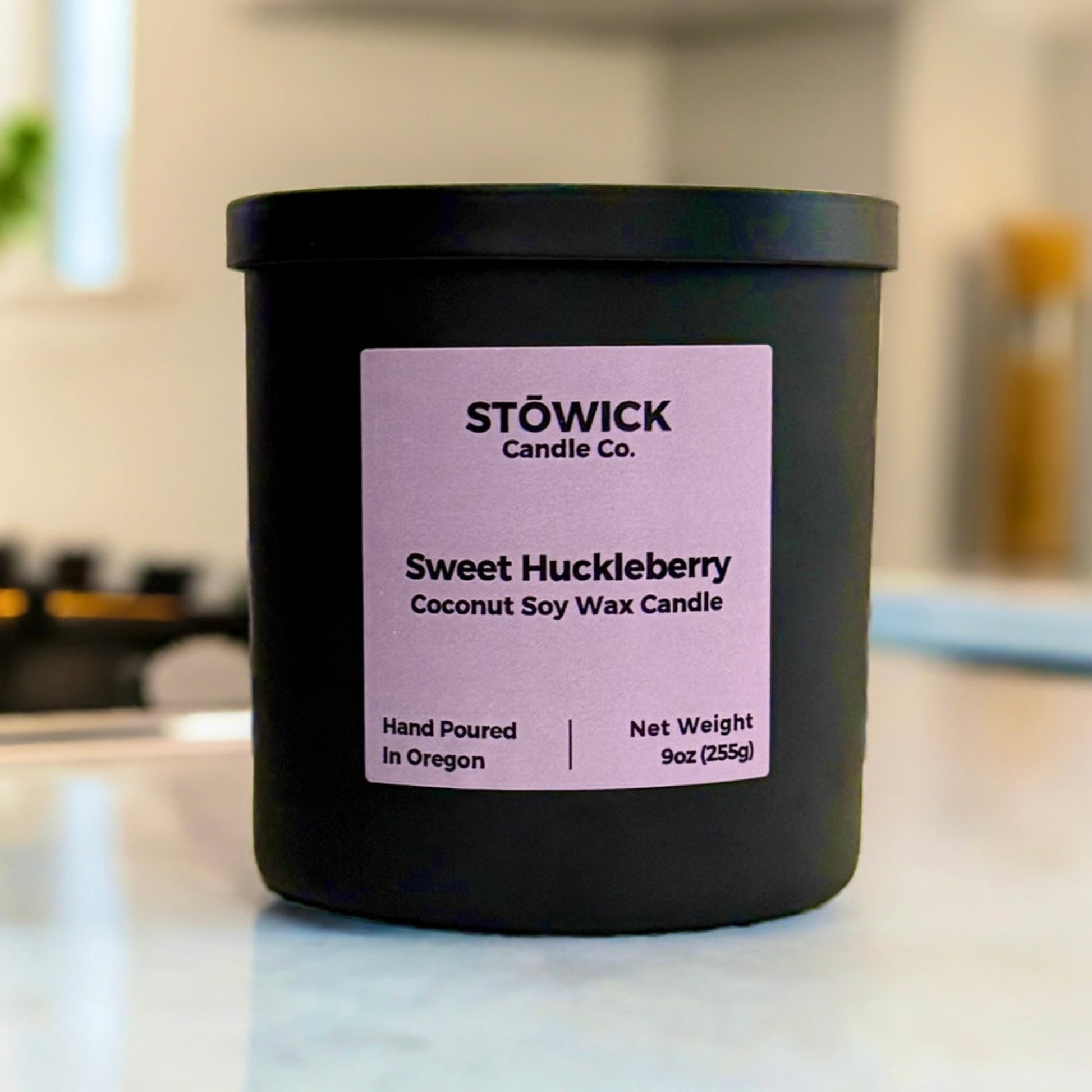 A 9oz sweet huckleberry tumbler candle. The candle has a black matte jar with a matching black lid. Sitting on a white kitchen counter with a purple label.