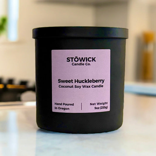 A 9oz sweet huckleberry tumbler candle. The candle has a black matte jar with a matching black lid. Sitting on a white kitchen counter with a purple label.
