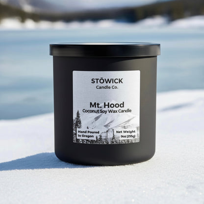 Mt. hood tumbler candle sitting in the snow in a black jar with a label featuring mt hood
