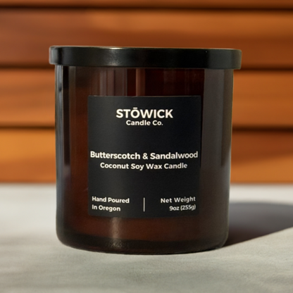 a 9 ounce butterscotch and sandalwood candle made from soy and coconut wax in an amber jar sitting on a white table wit a wood wall behind it