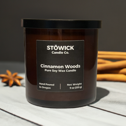 A 9 ounce cinnamon woods scented soy wax candle in an amber jar sitting on a table next to cinnamon sticks