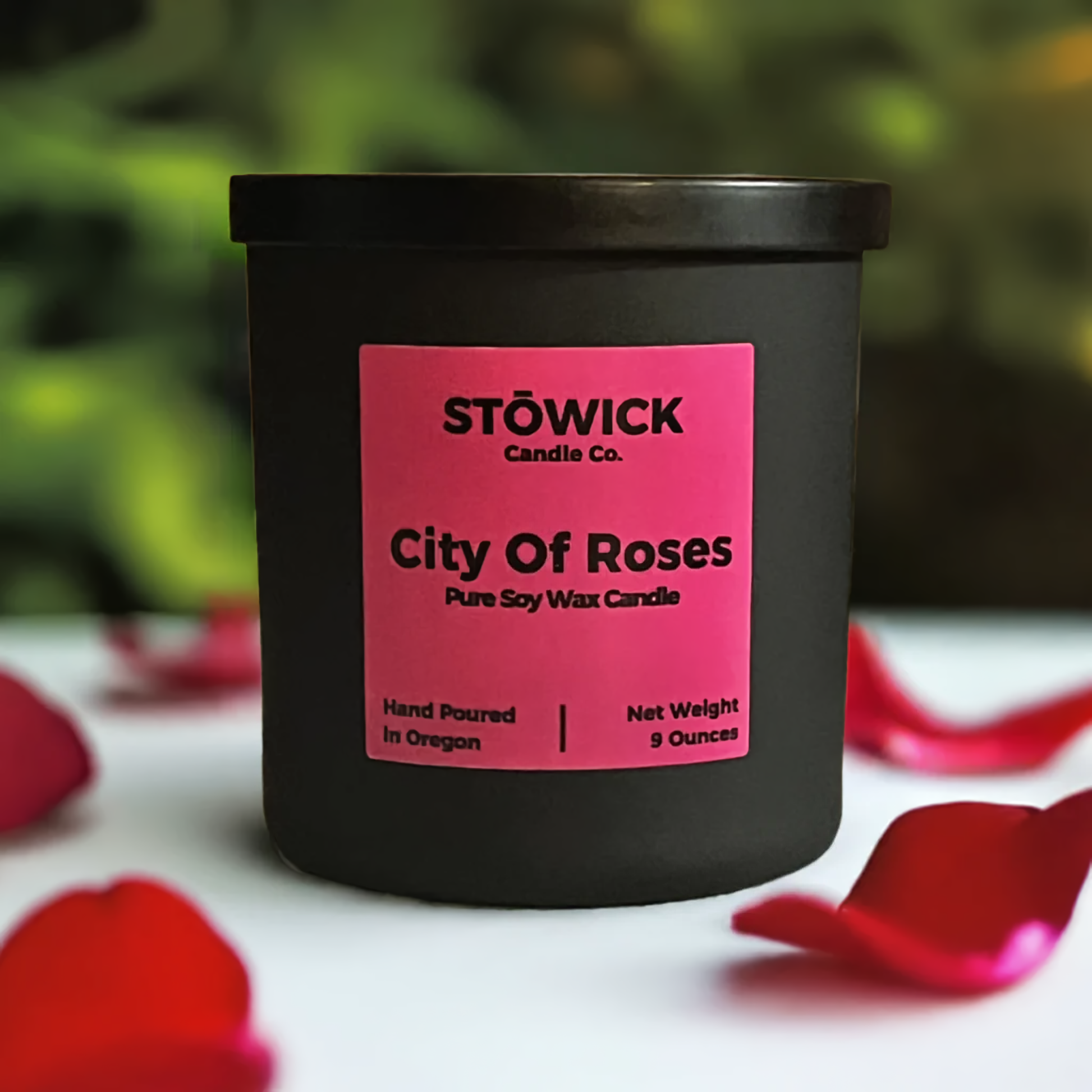 A city of roses scented soy wax candle in a black jar with a black lid and pink label sitting outside on a table next to rose petals