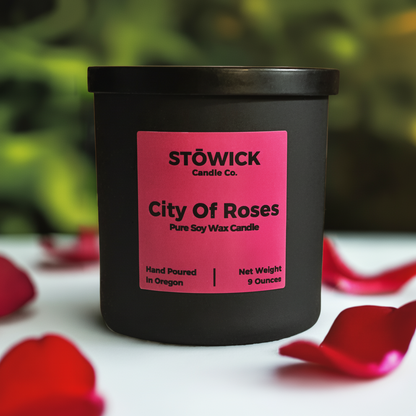 A city of roses scented soy wax candle in a black jar with a black lid and pink label sitting outside on a table next to rose petals