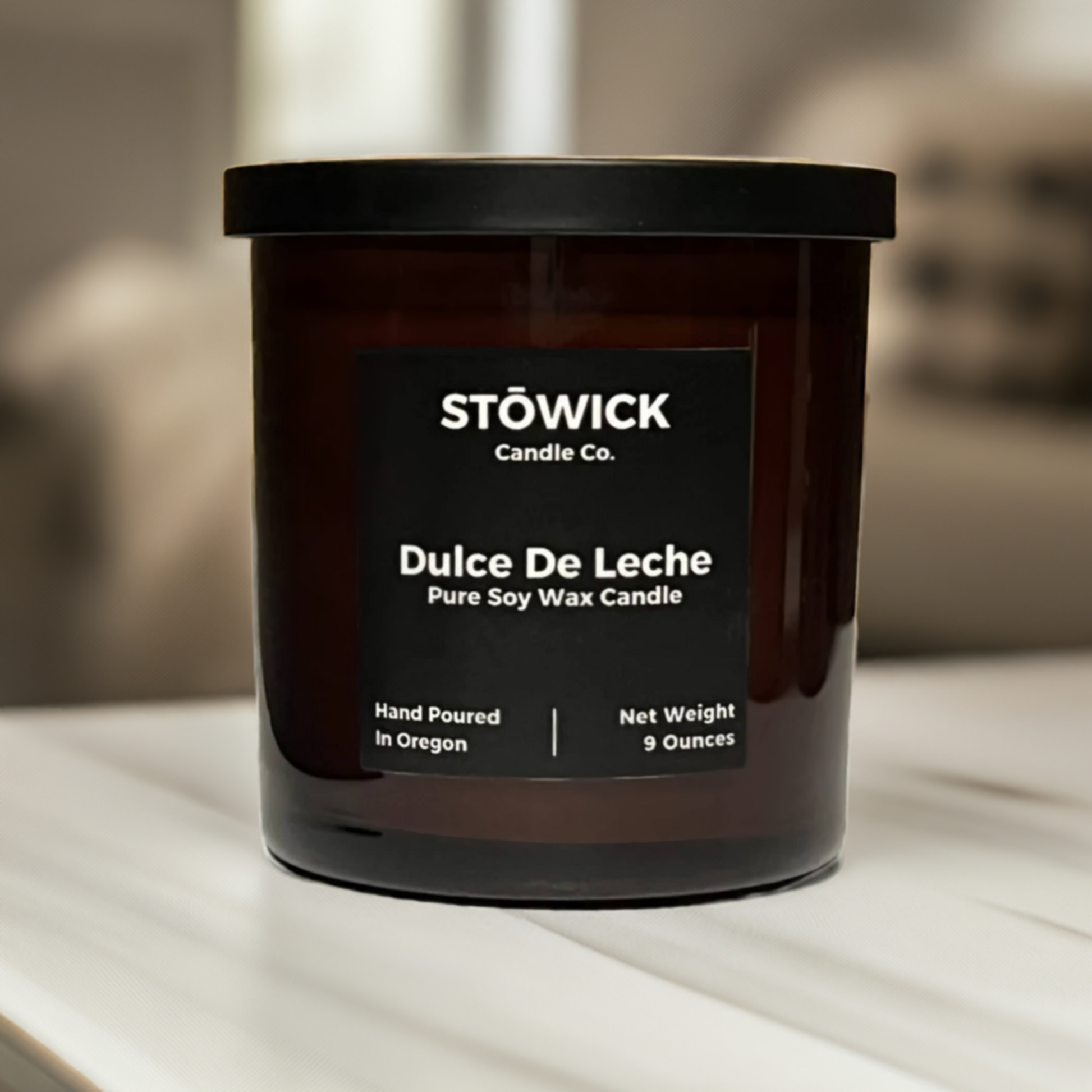 A dulce de leche scented candle in an amber jar with a black lid made from pure soy wax sitting on a white table