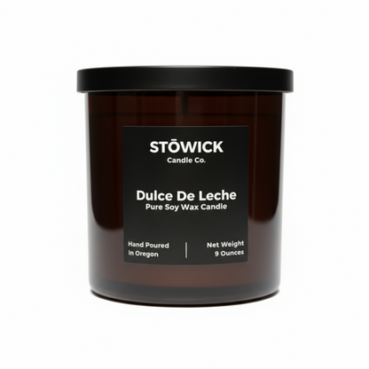 A dulce de leche scented candle in an amber jar with a black lid made from pure soy wax sitting on a white table