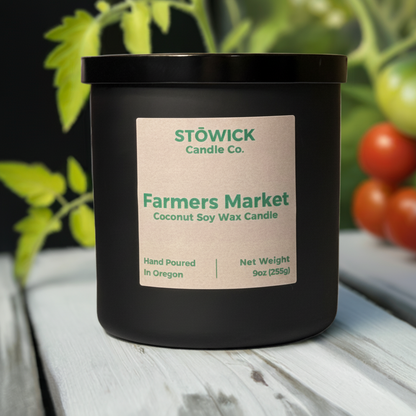 A farmers market scented candle in a 9 ounce black jar with a tan label sitting outside on a white wood table with tomato vines in the background