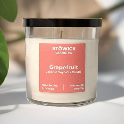 A grapefruit scented soy coconut wax candle in a clear jar with a pink label and a black lid on top of a table next to a grapefruit plant