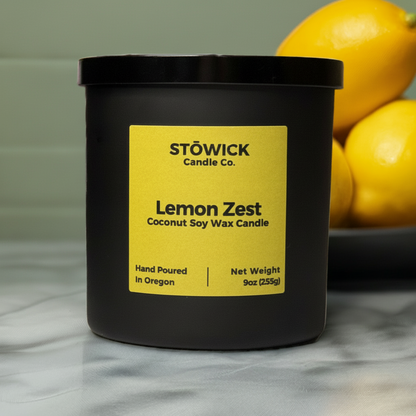 A 9 ounce lemon zest scented soy coconut wax candle in a black matte jar with a yellow label sitting on a kitchen counter with lemons behind it.