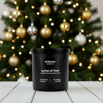 Black candle labeled 'Lump of Coal' by Stowick Candle Co. in front of a decorated Christmas tree.